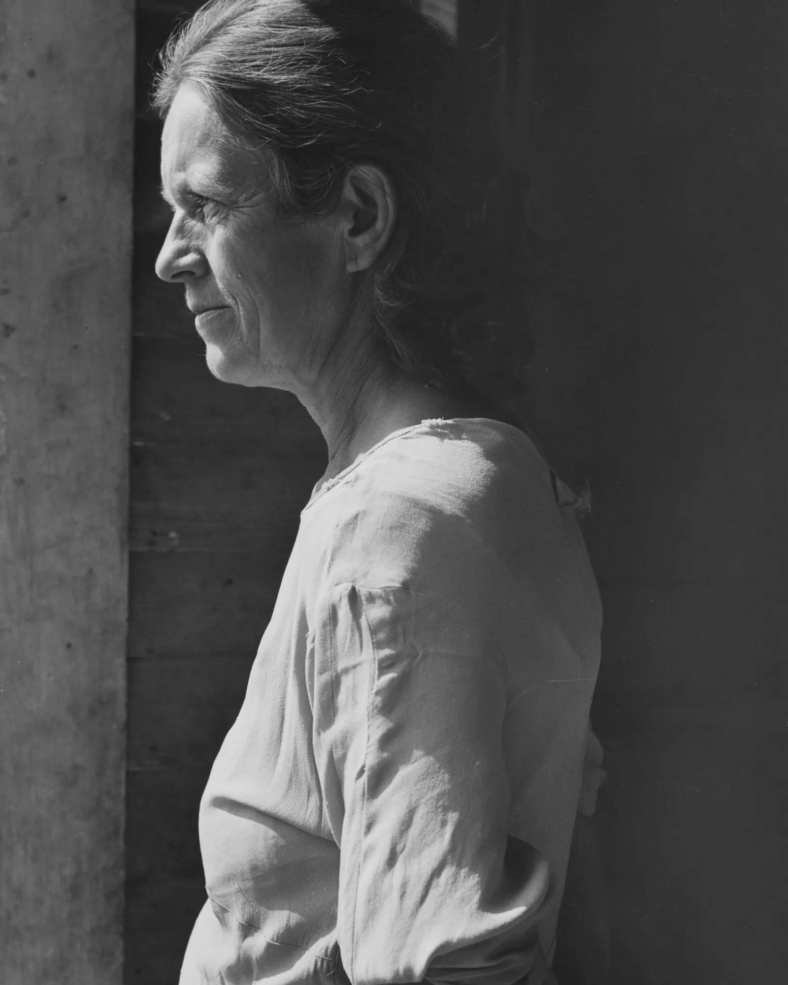 A woman standing in profile beside a window, lit from one side